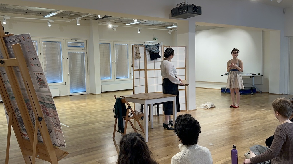 Two actors stand facing each other in mid-performance of 'Faith Hope Charity'. Both actors are standing  in front of a folding room divider in a bright acting studio. Other props in the scene include a table and chair and an easel propped to the left of the performers. A few students sit in the audience watching the performance.
